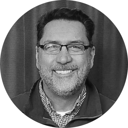 A black & white portrait of Bob Crimmins, a white man with a kind smile, looking straight on at the viewer. He has a beard, wears glasses, and is wearing a zip-up sweater over a checked button-down shirt.